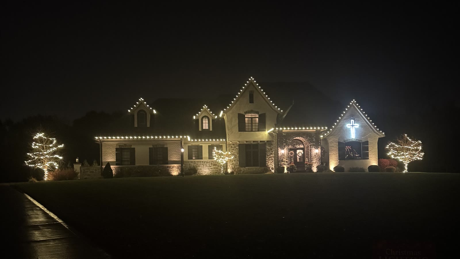 Large estate home lit with warm white Christmas lights along every roofline, a lit cross in the gable and two wrapped trees out front