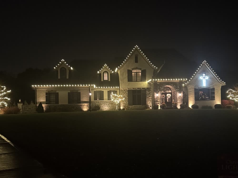 Large estate home with warm white lights along every roofline, a lit cross in the gable and two wrapped trees