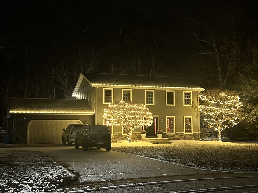 Two-story home with warm white roofline lights and Christmas lights wrapped around the front trees